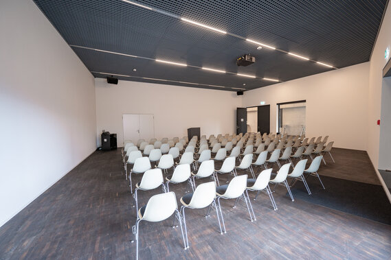 Ein Sitzungsraum mit Betonwänden und 10 Reihen Stühlen. Vor den Stühlen steht ein Rednerpult und ein Bild wird dahinter an die Wand projiziert. 
