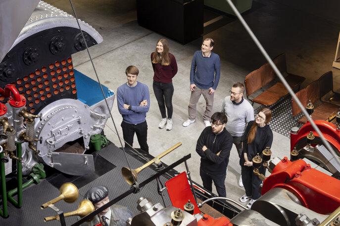 Personen stehen vor einem grossen Schiffsmotor in einer Ausstellung und werden von oben fotografiert.