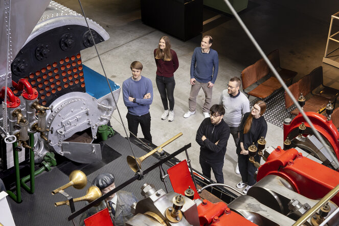 Personen stehen vor einem grossen Schiffsmotor in einer Ausstellung und werden von oben fotografiert. 