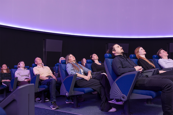 Eine Gruppe von Menschen entspannt sich in futuristischen Liegestühlen und blickt auf einen Kuppelbildschirm in einem schwach beleuchteten Planetarium.