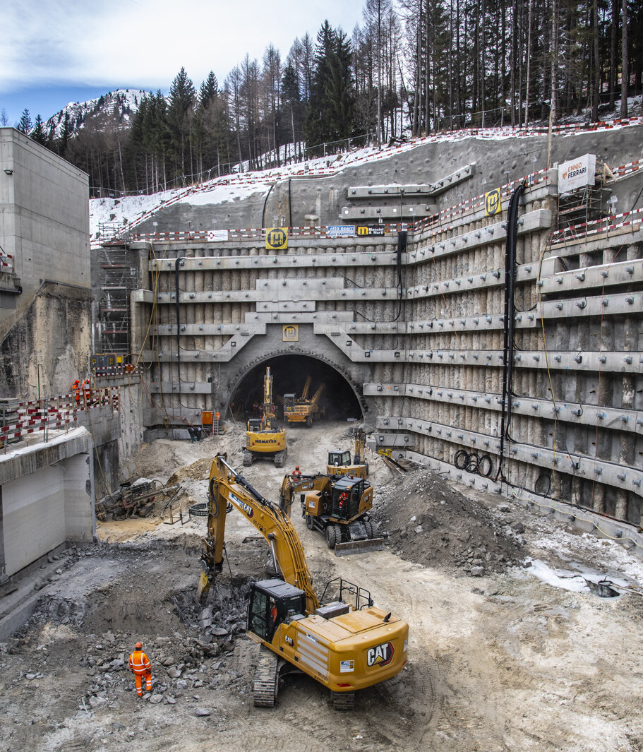 Baustelle mit schweren Maschinen, darunter Bagger, die an einem Tunnel arbeiten, der von steilem, schneebedecktem Gelände und Bäumen umgeben ist.
