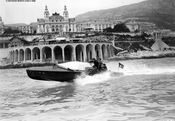 Ein altes Schwarz-Weiß-Foto von einem Schnellboot, das in der Nähe eines großen Casinos am Meer in Monaco um die Wette fährt und an dessen Rumpf Wellen plätschern.
