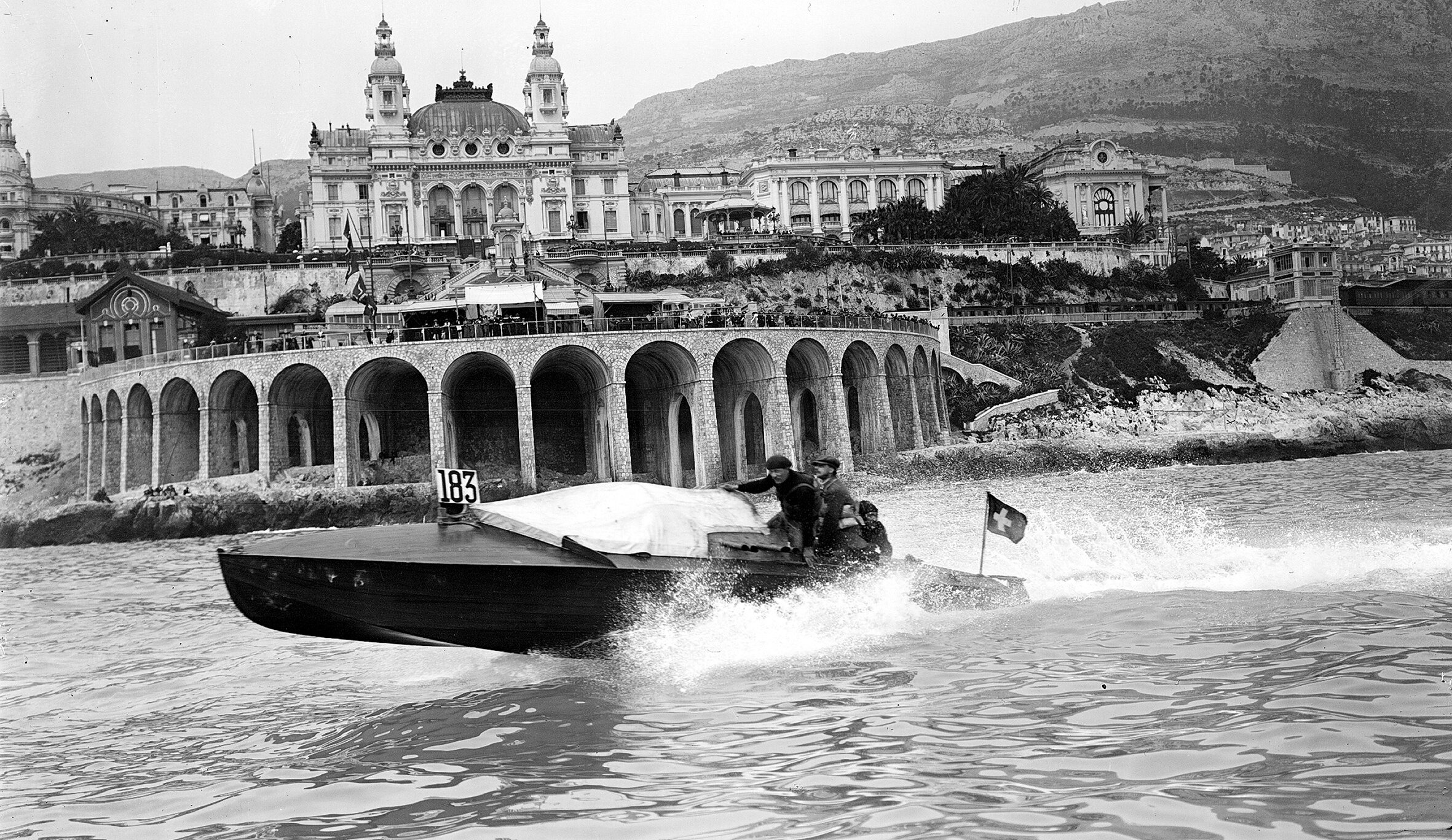 Ein altes Schwarz-Weiß-Foto von einem Schnellboot, das in der Nähe eines großen Casinos am Meer in Monaco um die Wette fährt und an dessen Rumpf Wellen plätschern.