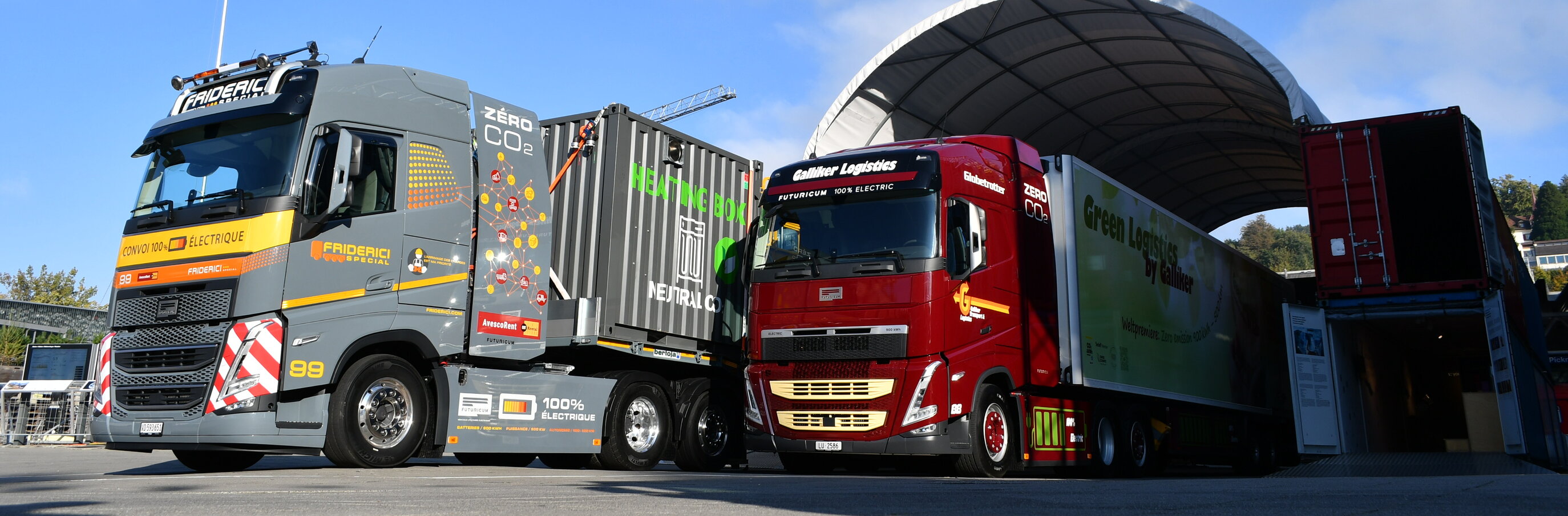 Zwei Elektro-Lkw, ein grauer und ein roter, parken unter einem Unterstand und demonstrieren umweltfreundliche Logistik und null CO2-Emissionen.