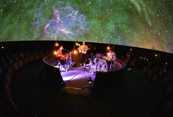 Eine Band tritt live unter einer schillernden Sternenprojektion in einem Planetarium auf, umgeben von einem Publikum in dunklen Sitzen.