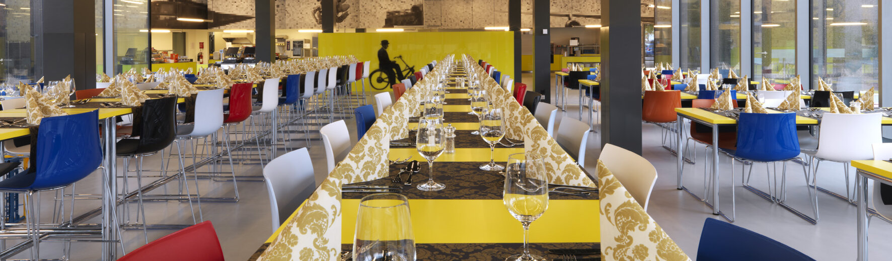 Lange, eckige Tische in einem Restaurant mit farbigen Stühlen und Servietten.