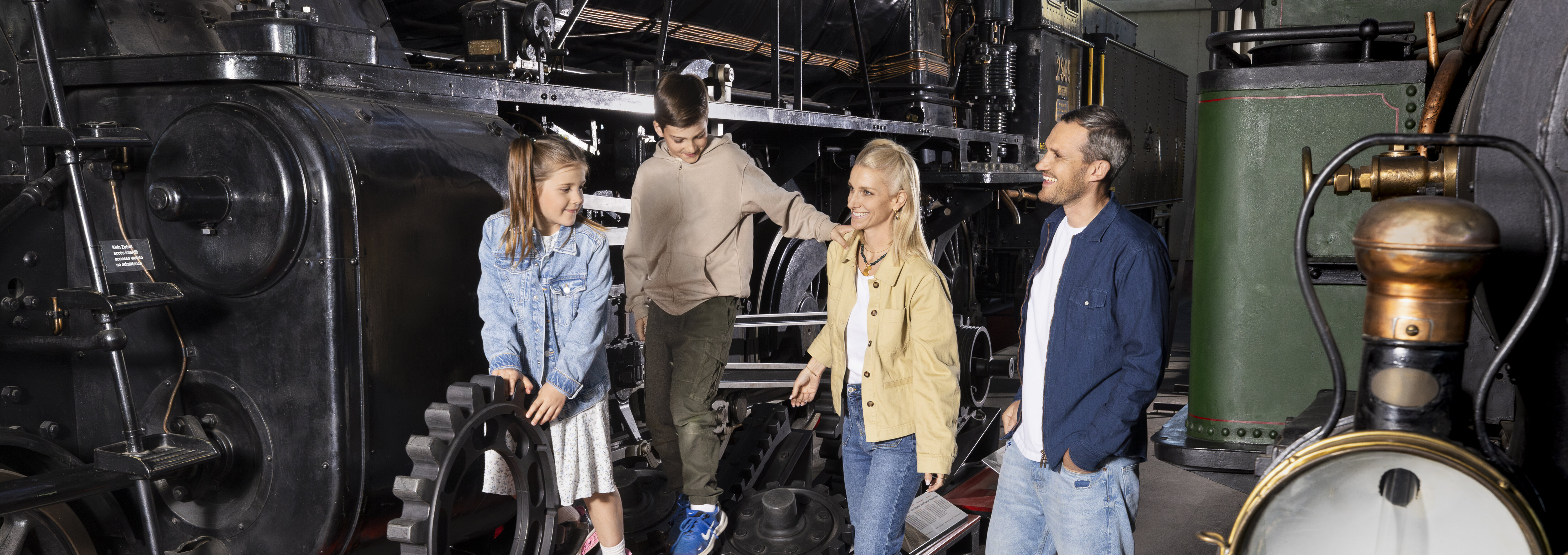 Eine vierköpfige Familie in der Schienenhalle im Verkehrshaus. Due Kinder klettern auf Zahnrädern herum, die Eltern stützen sie.