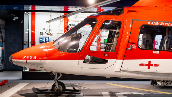 Ein rot-weisser Rega-Helikopter mit seinem Cockpit und den charakteristischen Markierungen ist vor einem informativen Ausstellungshintergrund ausgestellt.