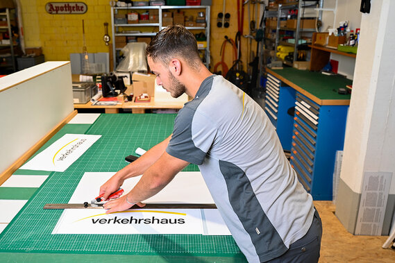 Ein Lehrling schneidet auf einer Werkbank in einer Werkstatt mit Lineal und Schere ein Schild mit der Aufschrift „verkehrshaus“ aus.