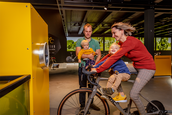 Eine Frau fährt mit einem Kind im Arm auf einem stationären Fahrrad, während ein anderer Erwachsener mit einem zweiten Kind in der Nähe steht, in einem lebendigen Ausstellungsraum.