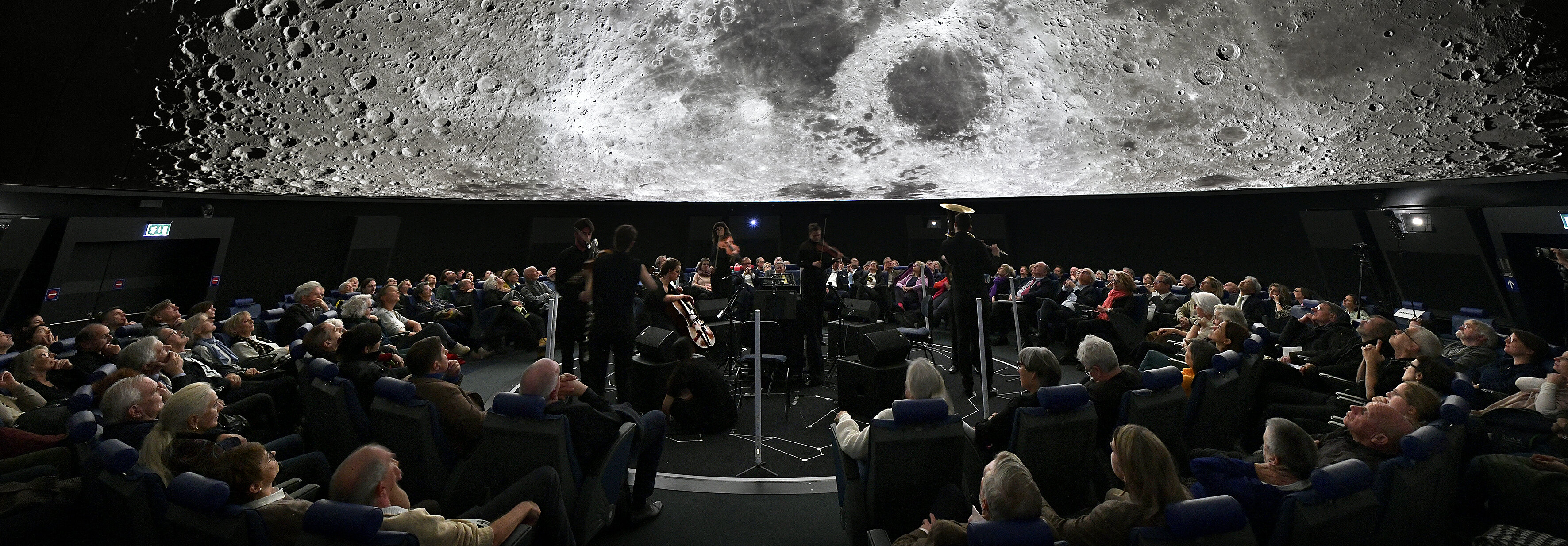 Ein großes Publikum verfolgt eine Vorstellung in einem Planetarium, in dem ein detailliertes Bild des Mondes über ihnen projiziert wird.