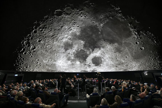 Ein großes Publikum verfolgt eine Vorstellung in einem Planetarium, in dem ein detailliertes Bild des Mondes über ihnen projiziert wird.