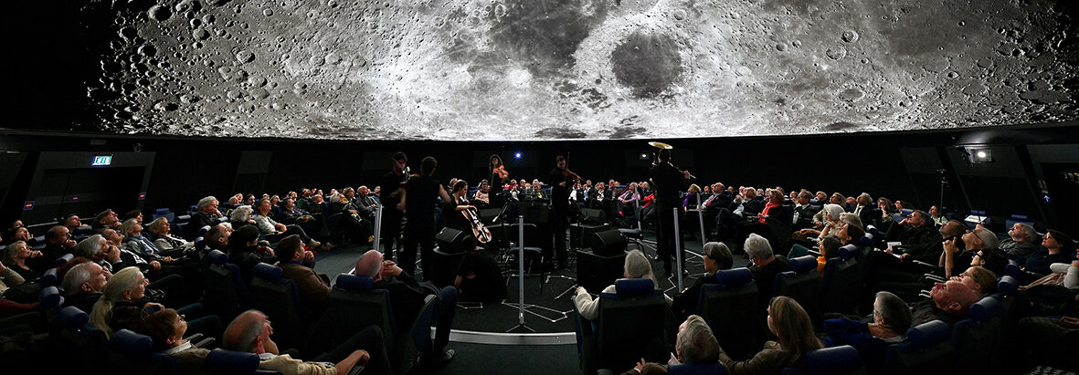 Ein großes Publikum verfolgt eine Vorstellung in einem Planetarium, in dem ein detailliertes Bild des Mondes über ihnen projiziert wird.