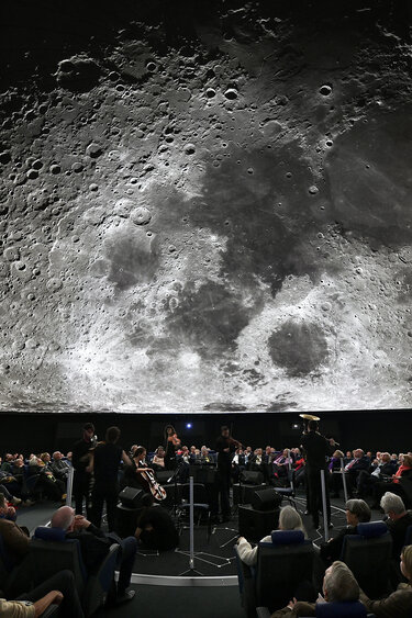 Ein großes Publikum verfolgt eine Vorstellung in einem Planetarium, in dem ein detailliertes Bild des Mondes über ihnen projiziert wird.