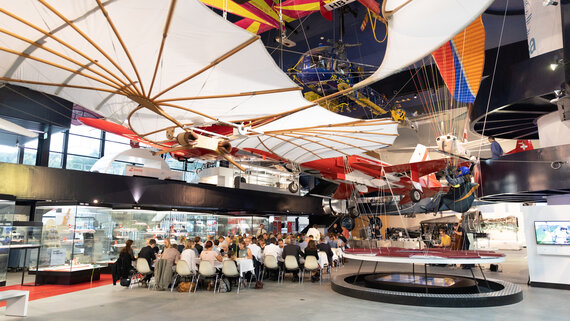 Leute sitzen an Tischen unter den Fliegern in der L&R Halle. 
