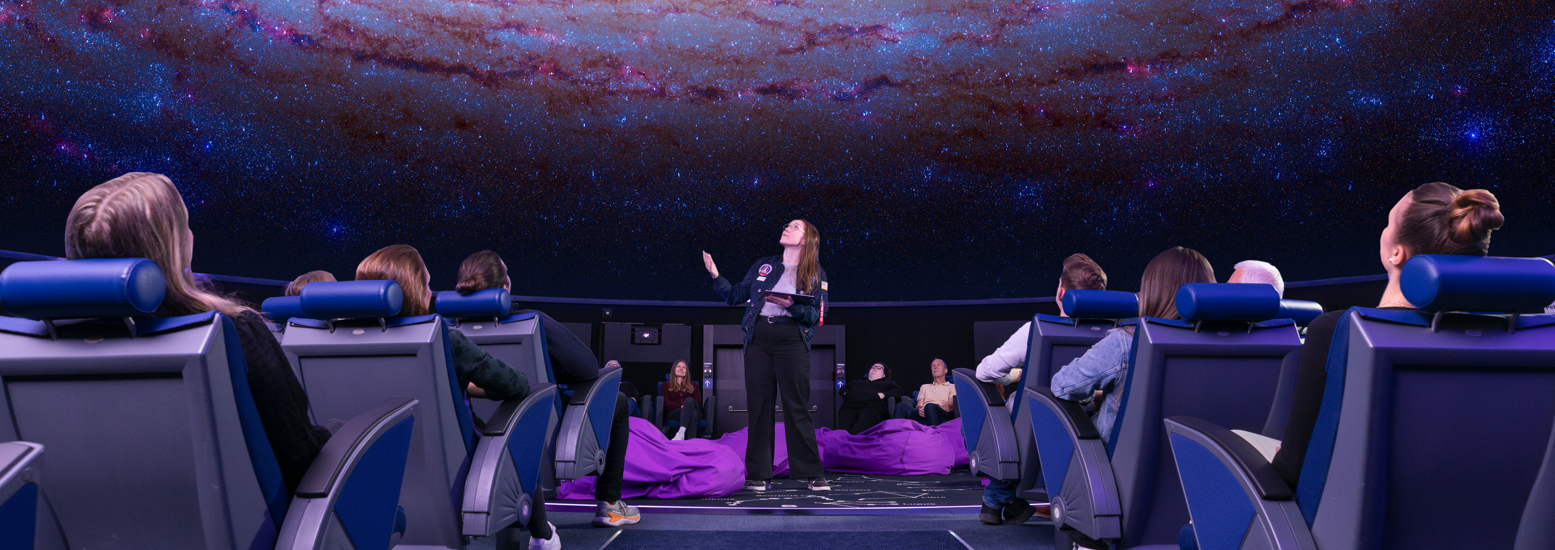 Eine Frau steht mit einem Tablet in der Mitte des Planetariums, über ihr ist das Universum zu sehen, um sie herum sitzen Zuschauer in Lehnstühle.