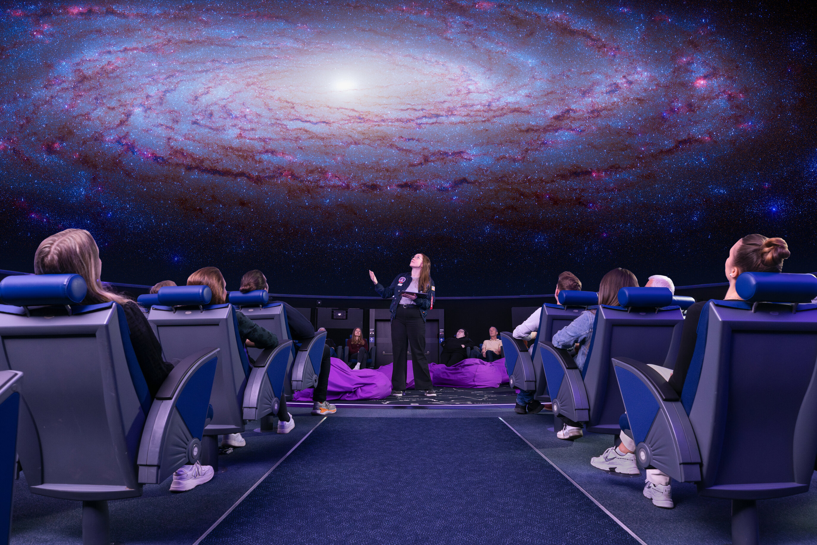 Eine Frau steht mit einem Tablet in der Mitte des Planetariums, über ihr ist das Universum zu sehen, um sie herum sitzen Zuschauer in Lehnstühle.