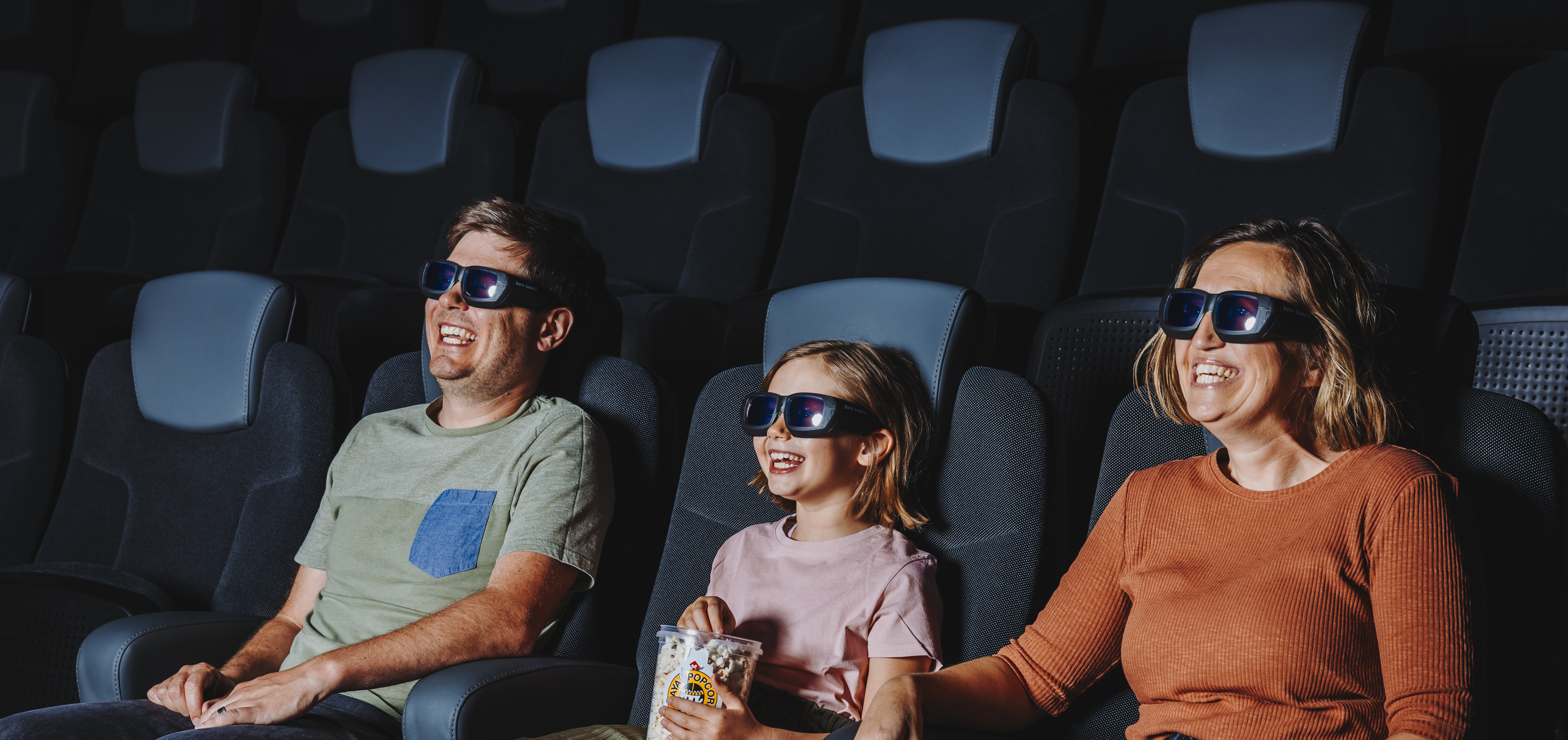 Eine Familie sitzt in bequemen Kinosesseln und schaut sich mit Popcorn in der Hand gemeinsam einen Film an.