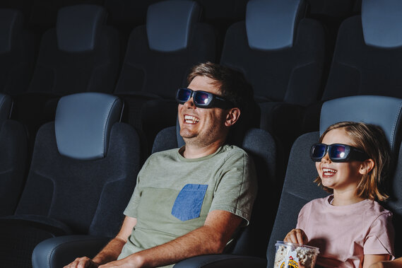 Eine Familie sitzt in bequemen Kinosesseln und schaut sich mit Popcorn in der Hand gemeinsam einen Film an.