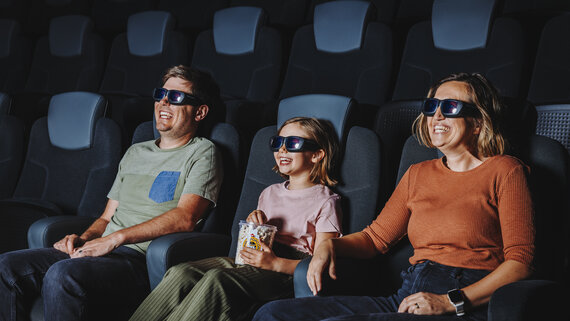 Eine Familie sitzt in bequemen Kinosesseln und schaut sich mit Popcorn in der Hand gemeinsam einen Film an.