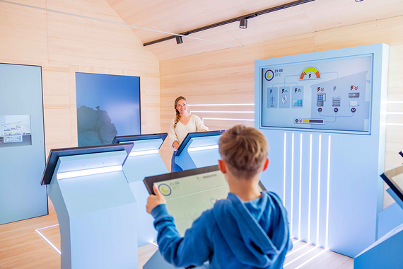 Zwei Kinder beschäftigen sich mit interaktiven Displays in einem modernen, mit Holz verkleideten Raum, in dem ein grosser Bildschirm Daten zum Energieverbrauch anzeigt.