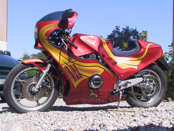 Ein rot-goldenes Custom-Motorrad, das auf einem Schotterplatz geparkt ist, präsentiert sich mit schnittigem Design und modernen Merkmalen unter einem klaren blauen Himmel.