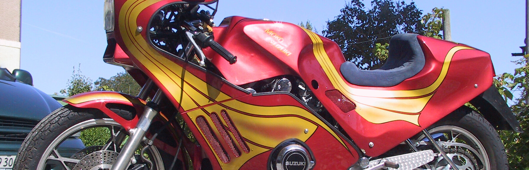 Ein rot-goldenes Custom-Motorrad, das auf einem Schotterplatz geparkt ist, präsentiert sich mit schnittigem Design und modernen Merkmalen unter einem klaren blauen Himmel.