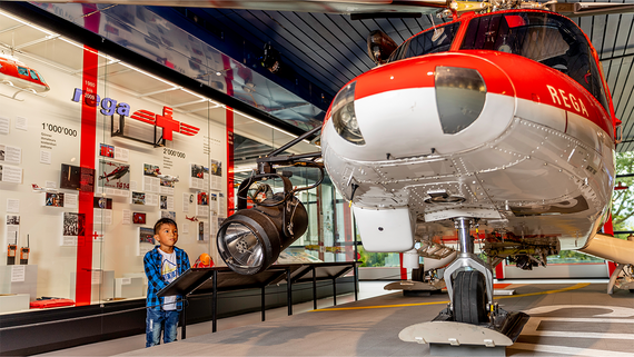 Ein Kind betrachtet einen ausgestellten rot-weißen Hubschrauber mit Informationstafeln und Artefakten über Rettungsdienste aus der Luft im Hintergrund.