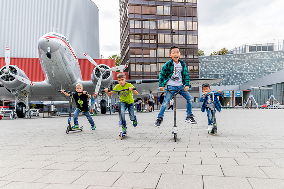  Eine Gruppe von Kindern fährt mit ihren Rollern vor einem alten Flugzeug und modernen Gebäuden.