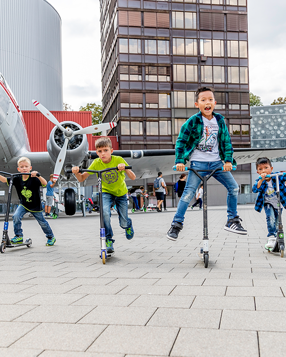  Eine Gruppe von Kindern fährt mit ihren Rollern vor einem alten Flugzeug und modernen Gebäuden.