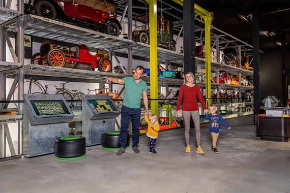Eine Familie erkundet eine Museumsausstellung mit Oldtimern und Fahrrädern und interaktiven Exponaten in einem modernen, großzügigen Ambiente.