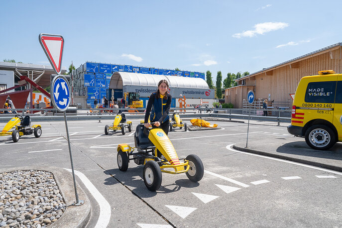 Ein Kind fährt in einem gelben Go-Kart auf einem Miniatur-Verkehrskurs, mit Straßenschildern und einem gelben Servicefahrzeug im Hintergrund.