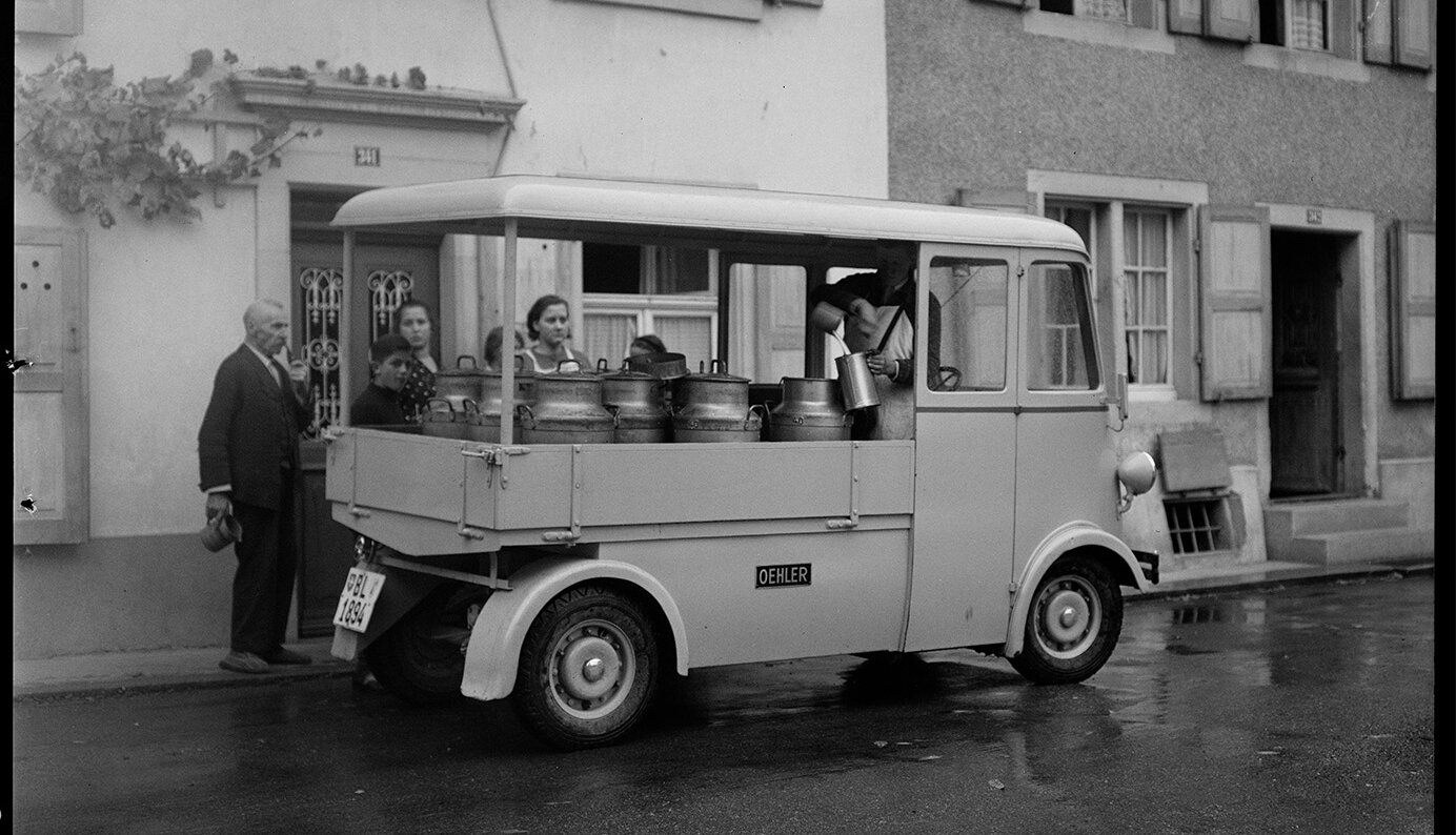 Ein kleiner, mit Metallbehältern gefüllter Oldtimer-Lieferwagen, der vor einem malerischen Gebäude mit hölzernen Fensterläden in einer nassen Straße geparkt ist.