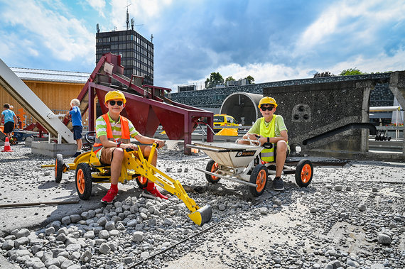 Zwei Kinder steuern Miniatur-Baufahrzeuge auf einem Schotterplatz, umgeben von Baumaschinen und einem bewölkten Himmel.