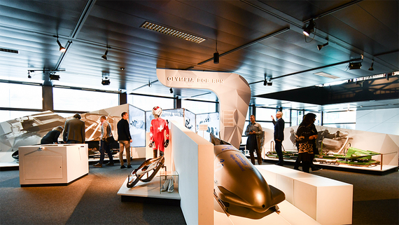 Innenansicht einer Museumsausstellung mit olympischen Bobschlitten, Rennanzügen und Besuchern, die die Ausstellungsstücke erkunden.
