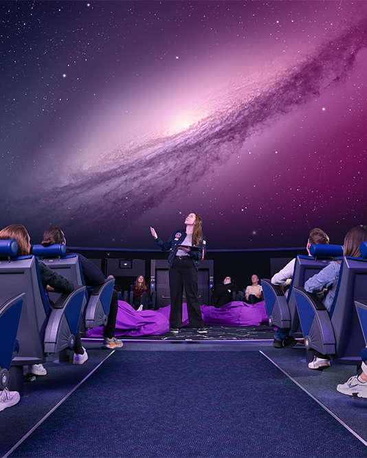 Ein Moderator unterhält das Publikum unter einer galaktischen Kuppel in einem Planetarium, in der Sterne und kosmische Bilder zu sehen sind.
