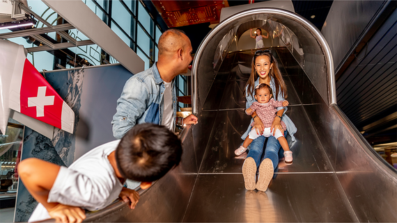 Eine Familie geniesst eine lustige Rutsche in einem Indoor-Spielbereich, umgeben von buntem Dekor und einer Schweizer Flagge im Hintergrund.