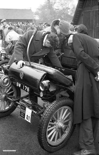 Eine Gruppe von Menschen betrachtet einen Oldsmobile von 1903 und konzentriert sich auf seine Mechanik bei einer Oldtimer-Veranstaltung.
