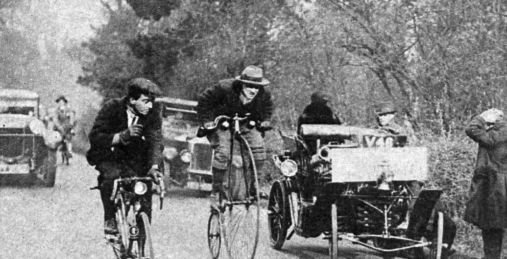 Eine alte Straßenszene mit einem Radfahrer auf einem Hochrad, einem anderen Radfahrer und frühen Autos inmitten von Bäumen.