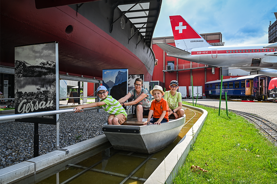 Drei Kinder spielen in einem kleinen bootähnlichen Gebilde vor dem Hintergrund alter Transportmittel, darunter Flugzeuge und Züge.
