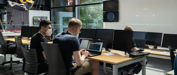 Drei Personen arbeiten an Schreibtischen mit mehreren Computermonitoren in einem modernen, von Tageslicht erhellten Büro.