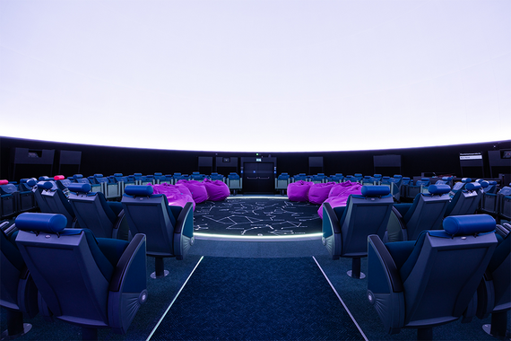 Innenraum eines Planetariums mit dunkelblauen Liegesitzen, rosafarbenen Sitzsäcken und einem kreisförmigen Lichtdesign auf dem Boden.