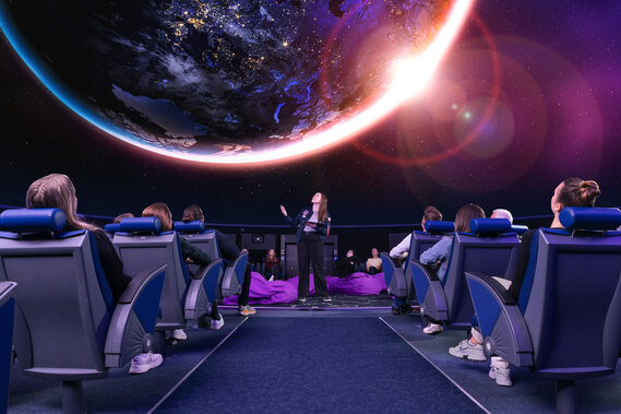 Eine Frau steht mit einem Tablet in der Mitte des Planetariums, über ihr ist die Erde zu sehen, um sie herum sitzen Zuschauer in Lehnstühle.
