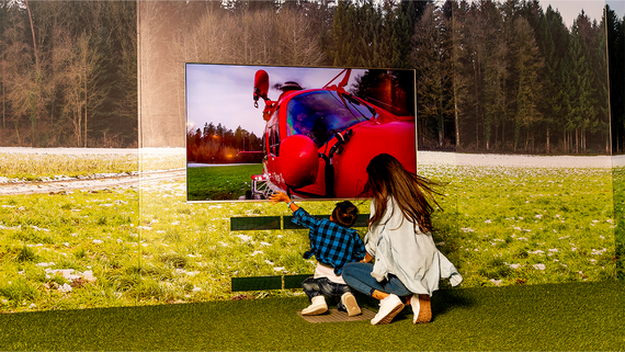 Eine Frau und ein Kind schauen auf einen grossen Fernseher, auf dem ein leuchtend roter Hubschrauber vor einer Kulisse aus Gras und Bäumen zu sehen ist.