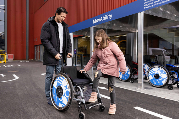 Ein Mann hilft einem Mädchen beim Einsteigen in einen Rollstuhl vor einem Geschäft für Mobilitätshilfen namens „MovAbility“, in dem verschiedene Rollstühle zu sehen sind.