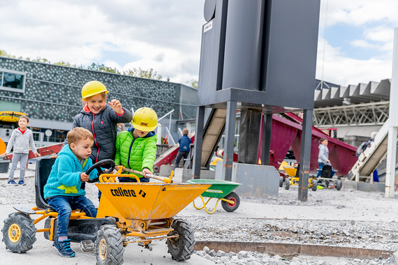 Drei Kinder mit Schutzhelmen spielen mit einem gelben Spielzeug-Baufahrzeug auf einem mit Kies gefüllten Spielplatz im Freien.