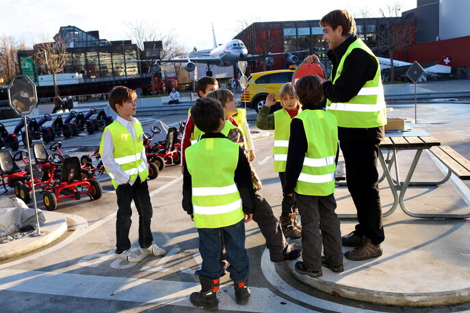 Fünf Kinder mit Leuchtwesten und eine Begleitperson mit Leuchtweste, sie stehen im Verkehrsgarten im Verkehrshaus.