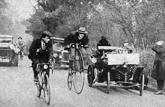 Ein Radfahrer und ein Mann auf einem Hochrad liefern sich ein Rennen auf einer Straße, flankiert von frühen Automobilen und einer zuschauenden Menge im Hintergrund.