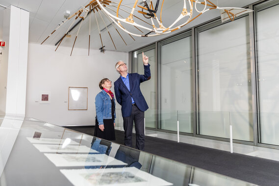 Ein Mann und eine Frau in einer Kunstausstellung. Sie begutachten ein aufgehängtes Objekt.