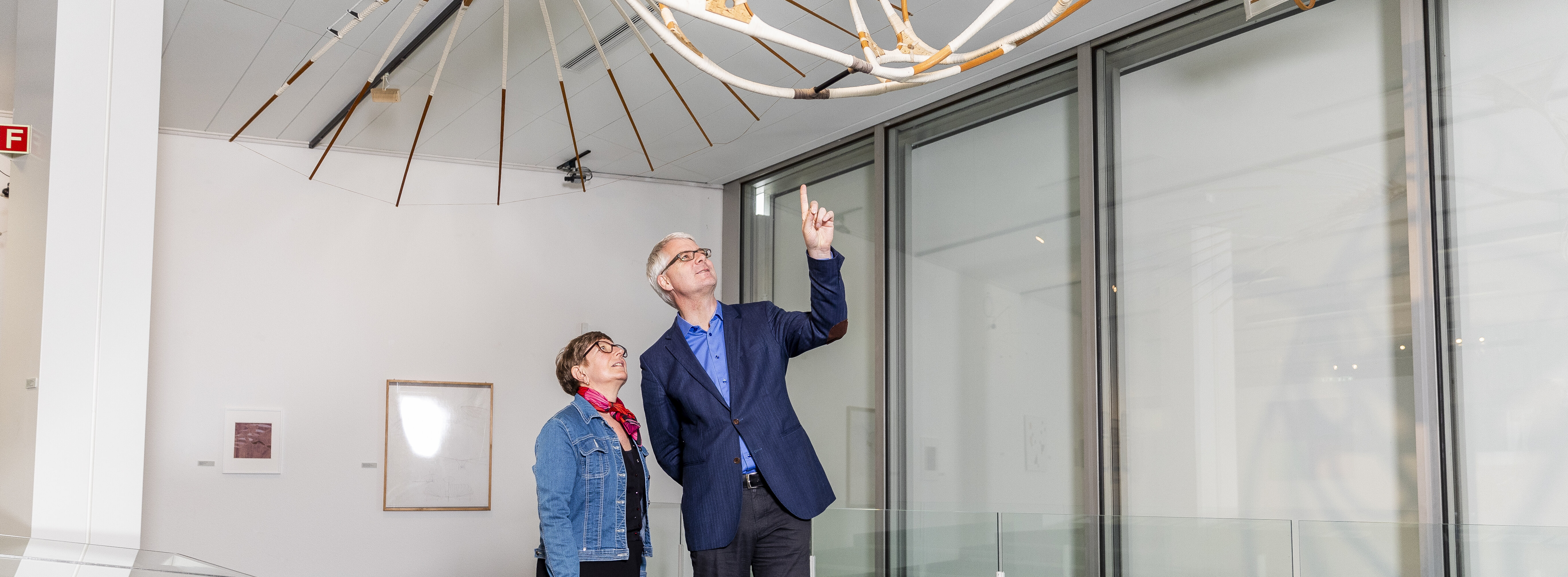 Ein Mann und eine Frau in einer Kunstausstellung. Sie begutachten ein aufgehängtes Objekt.